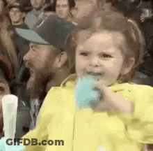A Little Girl In A Yellow Jacket Is Eating Cotton Candy While Sitting In A Crowd . GIF