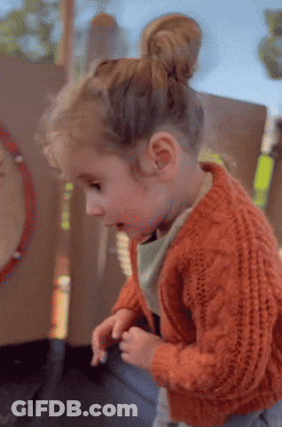 A Little Girl In An Orange Sweater Is Playing With Her Hands GIF