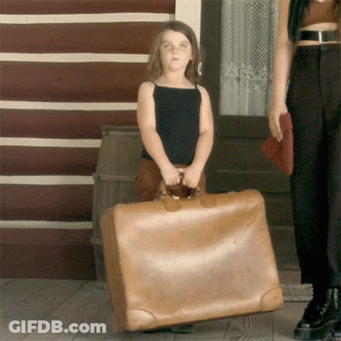 A Little Girl Is Holding A Large Brown Suitcase In Front Of A Log Cabin GIF