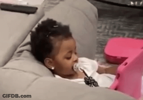 A Little Girl Is Laying On A Couch With A Pacifier In Her Mouth And Looking At A Tablet . GIF