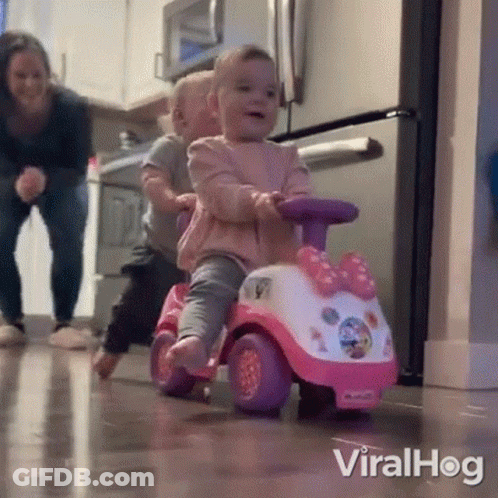 A Little Girl Is Riding A Pink And White Toy Car While A Little Boy Watches . GIF