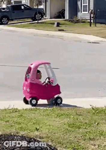A Little Girl Is Riding A Pink Toy Car Down A Street . GIF