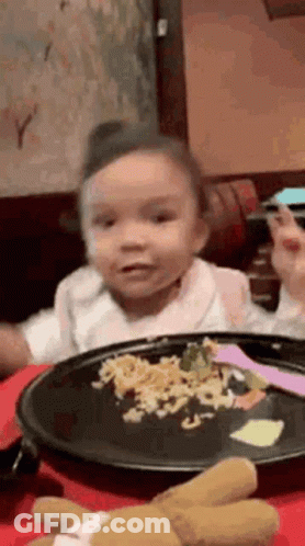 A Little Girl Is Sitting At A Table Eating A Plate Of Food . GIF