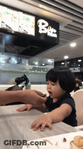 A Little Girl Is Sitting At A Table In Front Of A Sign That Says Bee Mian GIF