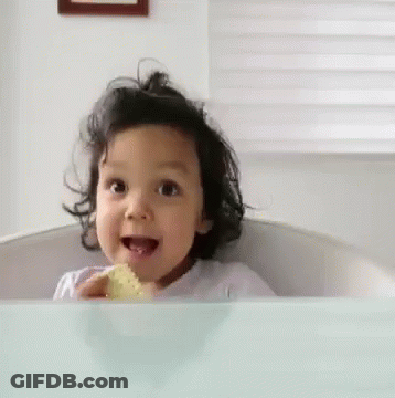 A Little Girl Is Sitting At A Table With A Piece Of Food In Her Hand And Smiling . GIF