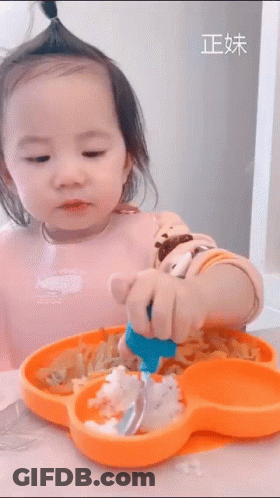 A Little Girl Is Sitting At A Table With A Plate Of Food And A Spoon In Her Hand . GIF