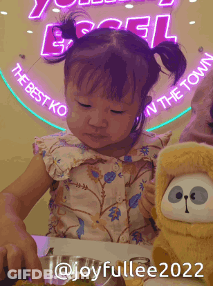 A Little Girl Is Sitting At A Table With A Stuffed Animal In Front Of A Sign That Says The Best Ko In The Town GIF