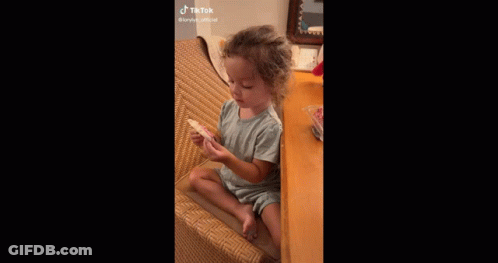 A Little Girl Is Sitting In A Chair Eating A Cookie . GIF