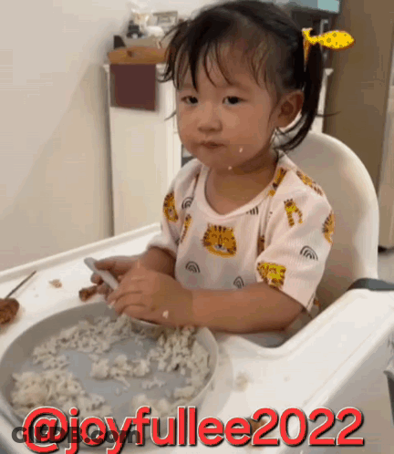 A Little Girl Is Sitting In A High Chair With A Plate Of Food And The Words Joyfullee2022 On The Bottom GIF