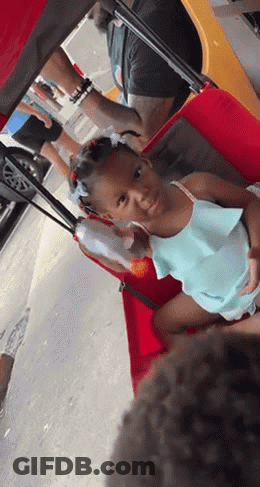 A Little Girl Is Sitting In A Red Cart With A Man Behind Her GIF