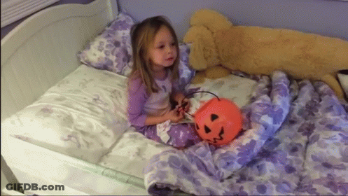 A Little Girl Is Sitting On A Bed Holding A Pumpkin Bucket GIF