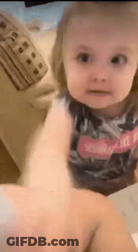 A Little Girl Is Sitting On A Couch Looking At The Camera While A Person Holds Her Hand . GIF
