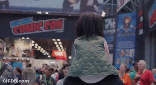 A Little Girl Is Sitting On A Person 'S Shoulders In Front Of A Sign That Says Comic Con GIF
