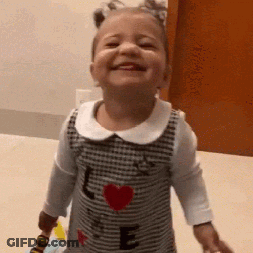 A Little Girl Is Smiling While Holding A Toy And Wearing A Dress With A Heart On It . GIF