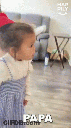 A Little Girl Is Standing In A Living Room Wearing A Red Hat And A Blue Dress And Saying Papa . GIF