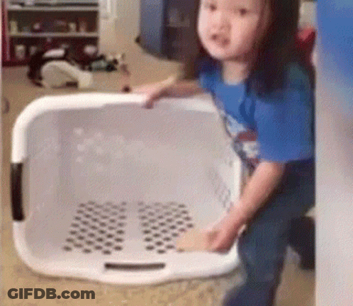 A Little Girl Is Standing Next To A Laundry Basket In A Room . GIF