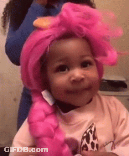 A Little Girl Is Wearing A Pink Wig And Smiling While A Woman Brushes Her Hair . GIF