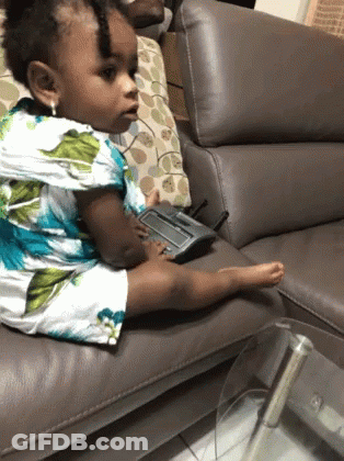 A Little Girl Sits On A Couch With A Remote Control In Her Hand GIF