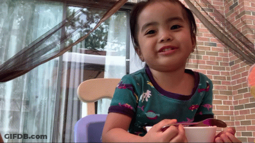 A Little Girl Sitting In A Chair With A Bowl Of Food And A Spoon GIF