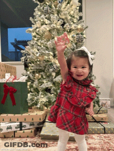 A Little Girl Stands In Front Of A Christmas Tree With Her Hand In The Air GIF