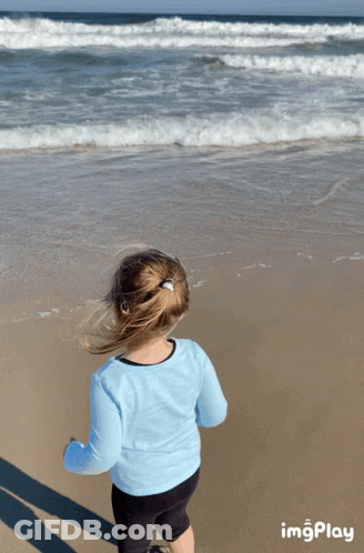A Little Girl Stands On A Beach Looking At The Ocean With Imgplay At The Bottom GIF