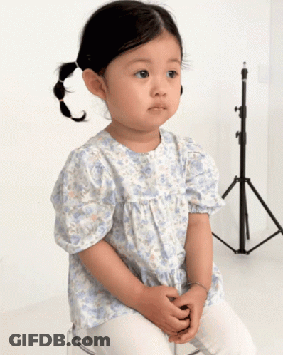 A Little Girl Wearing A Blue And White Floral Shirt Sits On A Clear Stool GIF