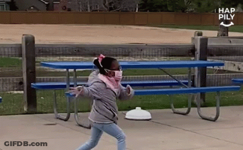 A Little Girl Wearing A Face Mask Is Walking In Front Of A Picnic Table . GIF