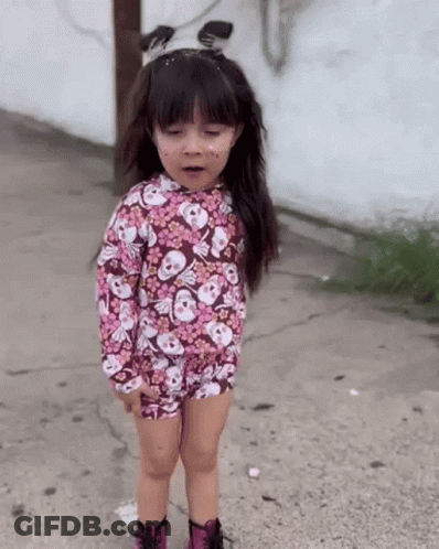 A Little Girl Wearing A Floral Shirt With Skulls On It Is Standing On A Sidewalk GIF