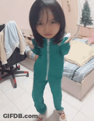 A Little Girl Wearing A Green Jumpsuit Is Standing In A Bedroom GIF