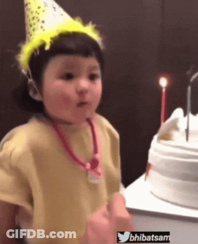 A Little Girl Wearing A Party Hat Stands In Front Of A Birthday Cake With Candles GIF