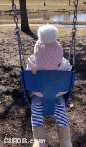 A Little Girl Wearing A Pink Hat And Striped Pants Is Sitting On A Blue Swing GIF