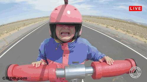 A Little Girl Wearing A Pink Helmet And A Blue Jacket Is Riding A Red Pipe On A Road With A Live Sign Above GIF