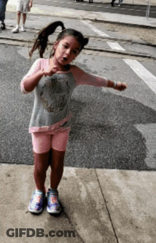 A Little Girl Wearing A Skull Shirt And Shorts Is Standing On The Sidewalk GIF