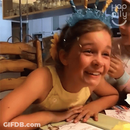 A Little Girl Wearing A Unicorn Headband Smiles While Sitting At A Table . GIF