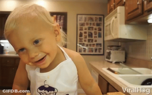 A Little Girl Wearing An Apron With A Cupcake On It Smiles In The Kitchen GIF