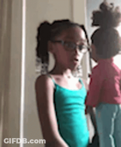 A Little Girl Wearing Glasses Is Standing Next To A Doll In A Room . GIF