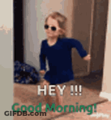 A Little Girl Wearing Sunglasses And A Blue Dress Is Standing In A Doorway And Saying Good Morning . GIF