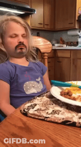 A Little Girl With A Fake Mustache Is Sitting At A Table With A Plate Of Food In Front Of Her GIF