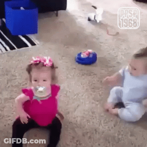 A Little Girl With A Pacifier In Her Mouth Is Sitting Next To A Baby . GIF