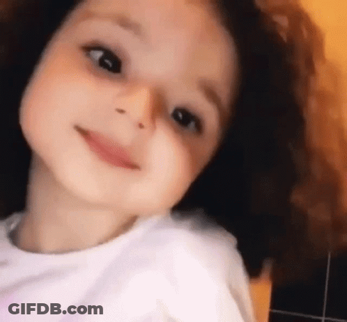 A Little Girl With Curly Hair Is Smiling And Looking Up At The Camera . GIF