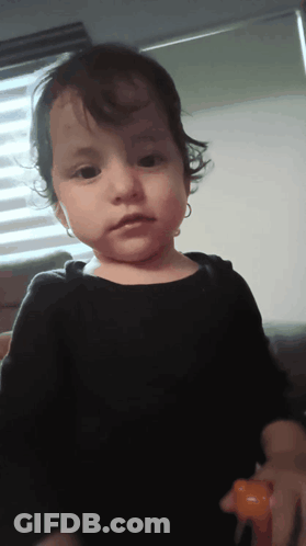 A Little Girl With Curly Hair Is Wearing A Black Shirt And Earrings GIF