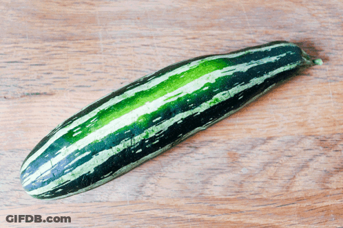 A Long Green And Black Striped Zucchini On A Wooden Table GIF