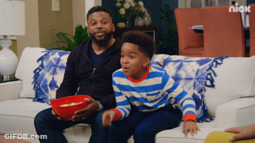 A Man And A Boy Are Sitting On A Couch With A Bowl Of Popcorn And A Nick Logo Behind Them GIF