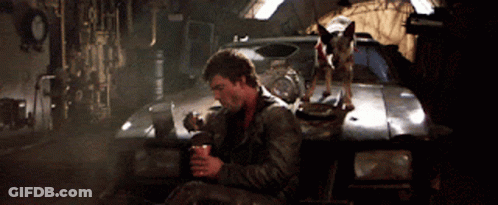 A Man And A Dog Are Sitting In Front Of A Car In A Garage GIF