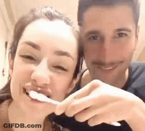 A Man And A Woman Are Brushing Their Teeth Together And Smiling For The Camera . GIF