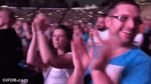 A Man And A Woman Are Clapping Their Hands In A Crowd . GIF