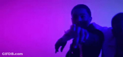 A Man And A Woman Are Dancing In Front Of A Purple Background . The Man Is Pointing At The Camera . GIF