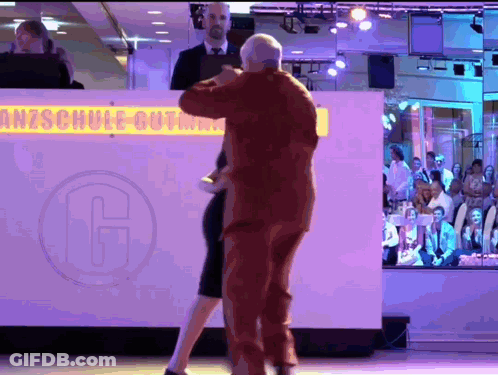 A Man And A Woman Are Dancing In Front Of A Sign That Says Anzschule GIF