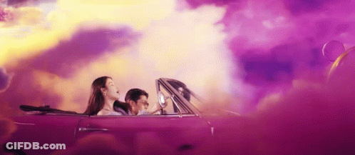 A Man And A Woman Are Driving A Purple Convertible Car . GIF