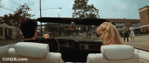 A Man And A Woman Are Driving Down A Street In A Car With A School In The Background GIF
